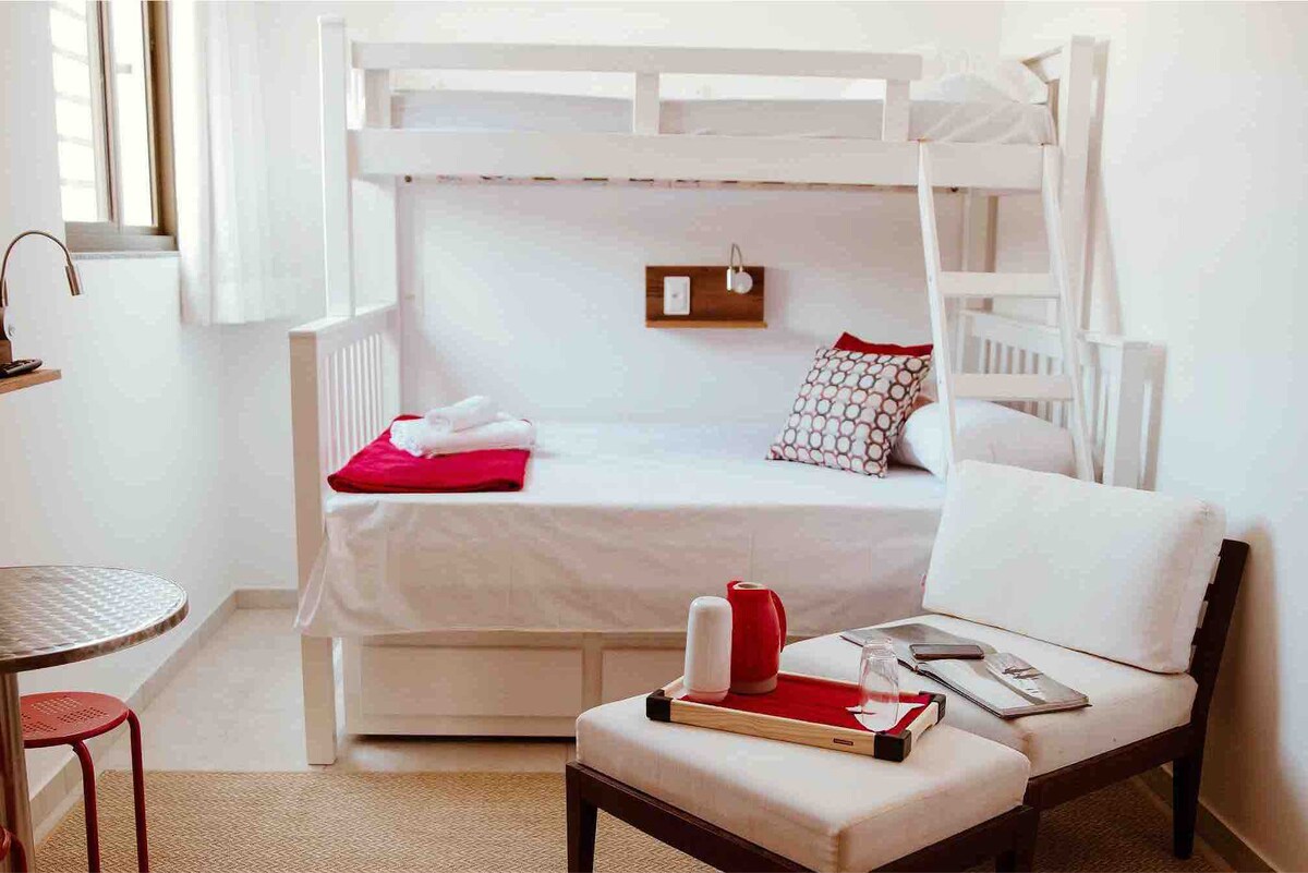 A bunk bed is positioned against the wall, featuring a single mattress on the bottom and a taller mattress above. A small bedside table with electrical outlets is mounted nearby. A cushioned chair and side table are placed in the corner, creating a cozy reading nook.