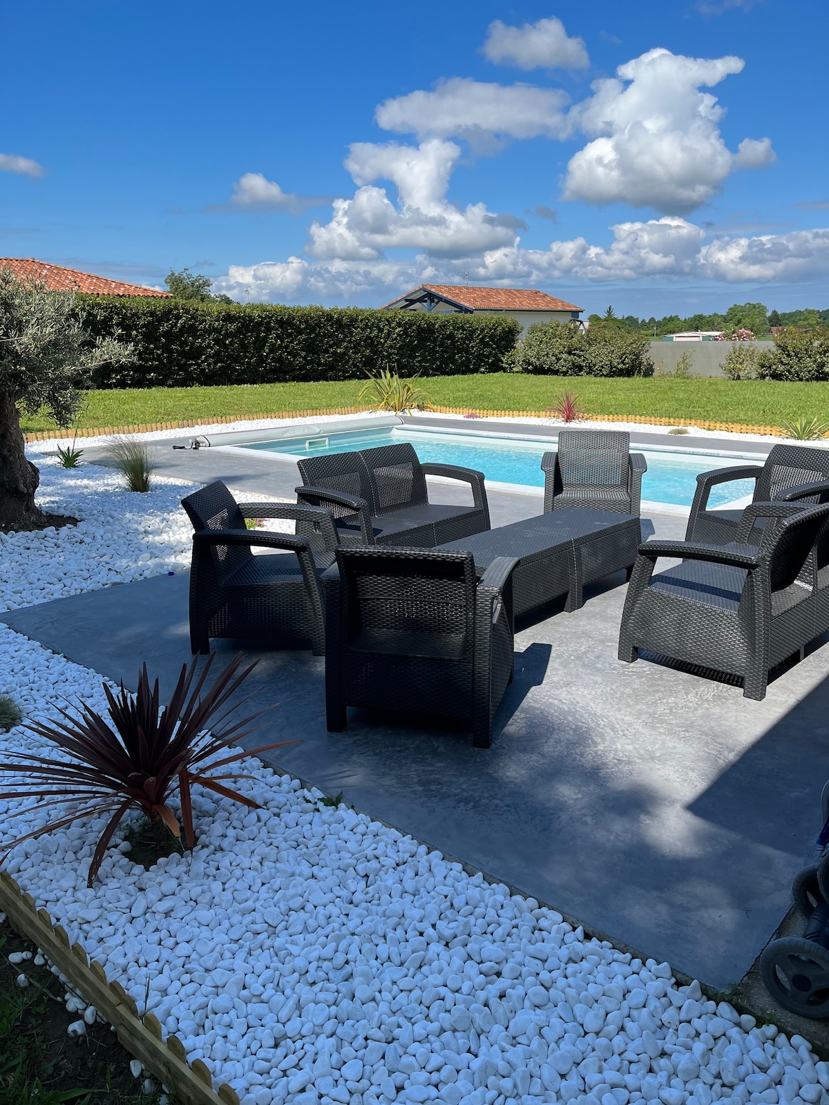 A serene outdoor area features a pool surrounded by tiled decking. Several black wicker chairs and a matching coffee table are arranged around the pool, inviting relaxation. Lush greenery and decorative white stones frame the space, enhancing the tranquil setting.