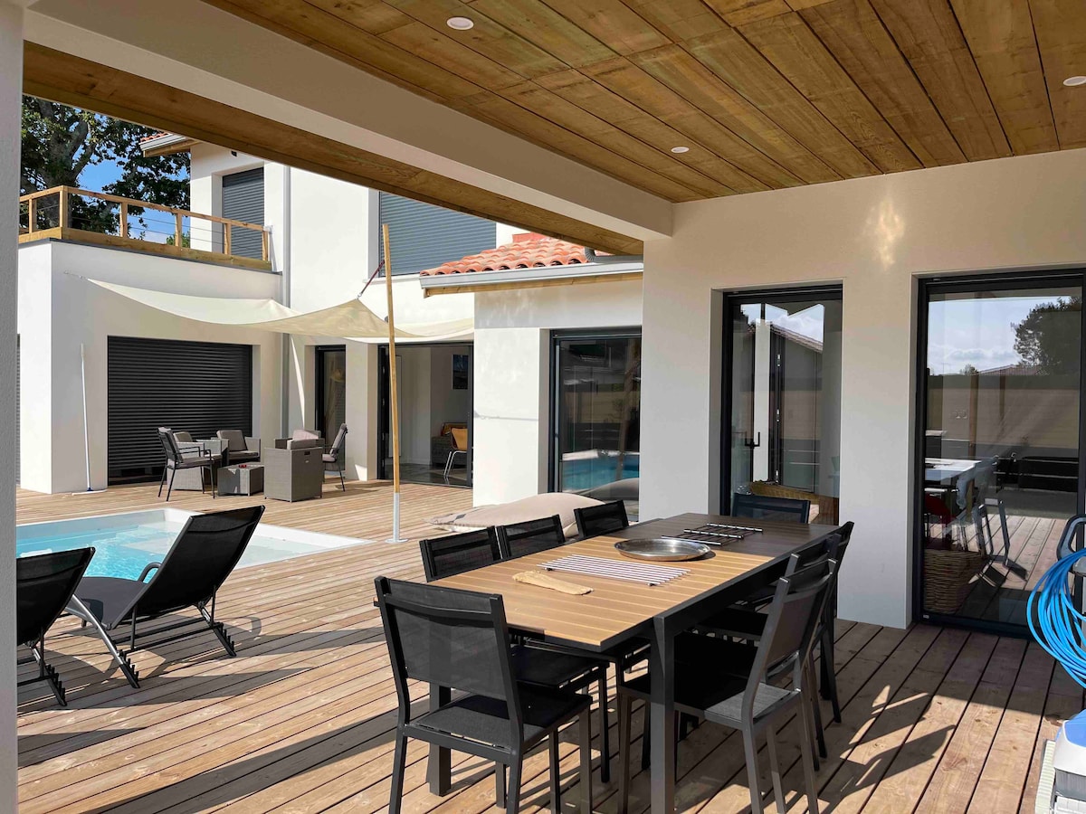 An outdoor dining area features a long wooden table with seating for eight, set beneath a covered terrace. The space overlooks a swimming pool surrounded by sun loungers, with views of the house's large glass doors opening to the interior.