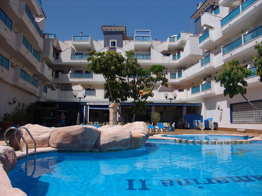 The image depicts a vibrant pool area surrounded by a multi-level residential building. Tropical plants and lounge chairs accompany the pool, which features a rock formation. Balconies with blue railings overlook the space, providing an inviting atmosphere for relaxation.