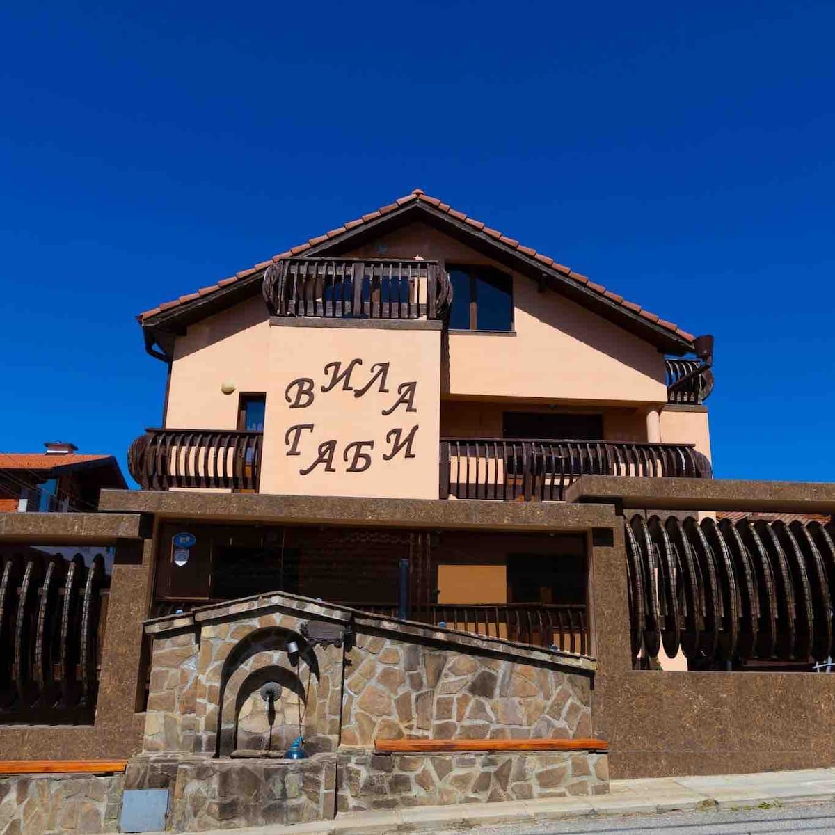 A three-story villa features a balanced brick and stucco exterior with a welcoming entrance. A stone wall surrounds the property, accented by a decorative fountain. Balconies with wooden railings enhance the second floor, while clear blue skies create a serene backdrop.