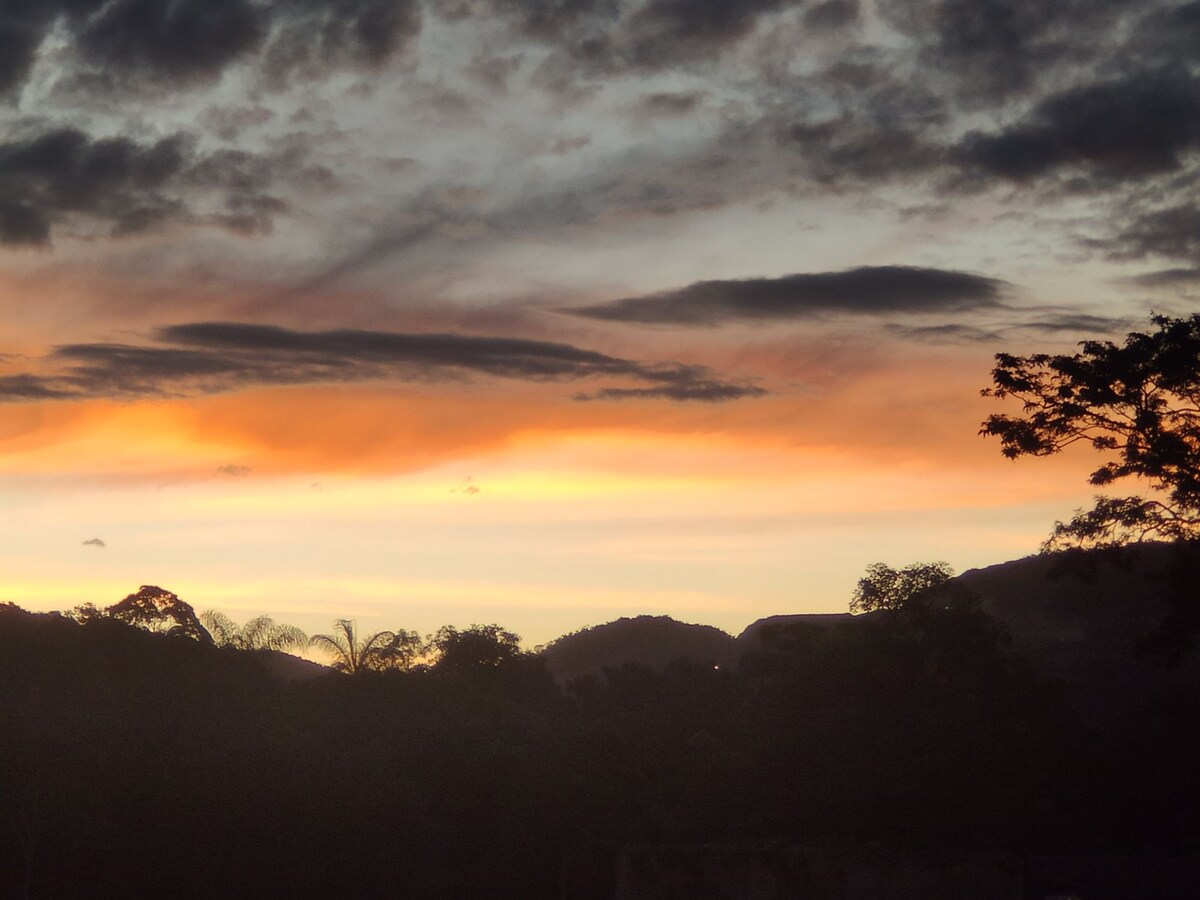 A sunset view displays vibrant hues of orange and purple across the sky, with silhouettes of distant mountains and trees framing the landscape. The scene captures the serene transition from day to night in a tranquil setting.