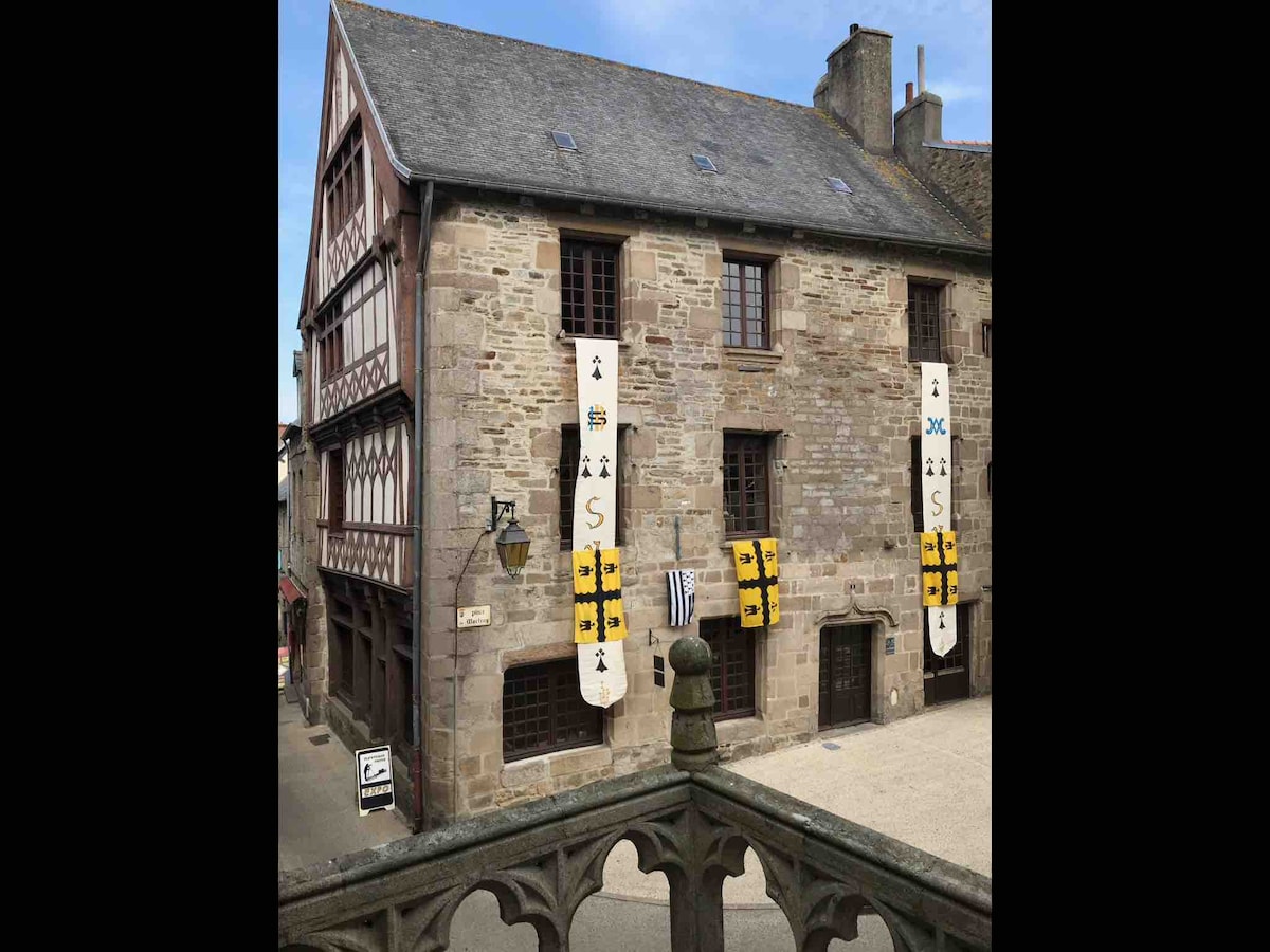 An historical building from the 15th century is presented, featuring a mix of stone and timber construction. Decorative banners in yellow and black adorn the facade. The entrance is framed by traditional architectural elements, with a central balcony providing a view of the surrounding area.
