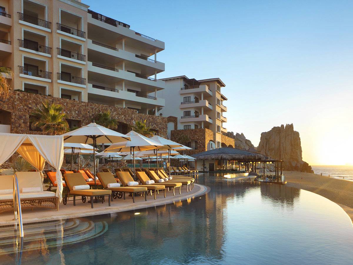 An expansive outdoor area is highlighted by lounge chairs under large umbrellas, positioned near a calm infinity pool. The water reflects the warm tones of the sunset, with resort buildings rising in the background, framed by large rock formations.
