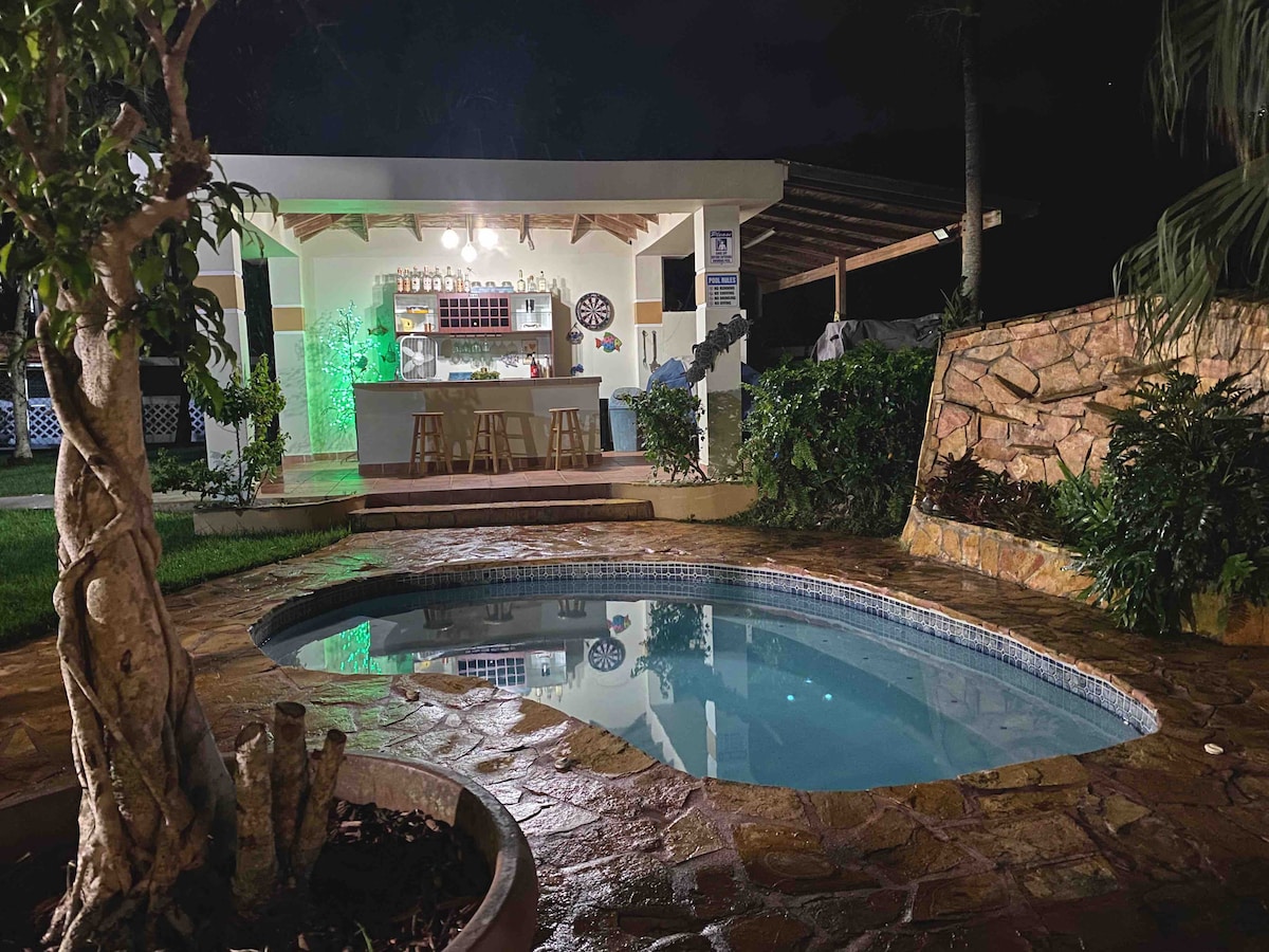 A private outdoor area features a serene pool with gently lit water, surrounded by tropical plants. An outdoor bar is visible under a covered patio, illuminated by soft lights. Stone pathways lead to the pool, complementing the natural ambiance of the space.