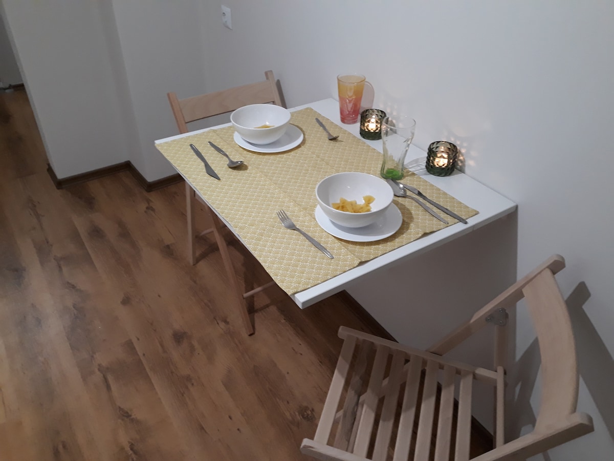 A simple dining table is set for two, featuring bowls and cutlery on a yellow placemat. Wooden folding chairs are positioned beside the table, adding to the minimalist decor. Soft candlelight creates a warm ambiance, enhancing the inviting presentation of the meal setup.