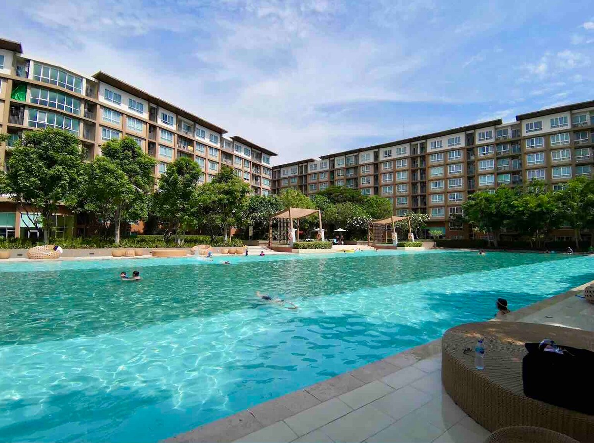 A large swimming pool is surrounded by well-maintained landscaping and multiple buildings. Lounge chairs are positioned along the pool's edges, with various guests enjoying the water. Clear blue skies and a few clouds enhance the serene atmosphere.