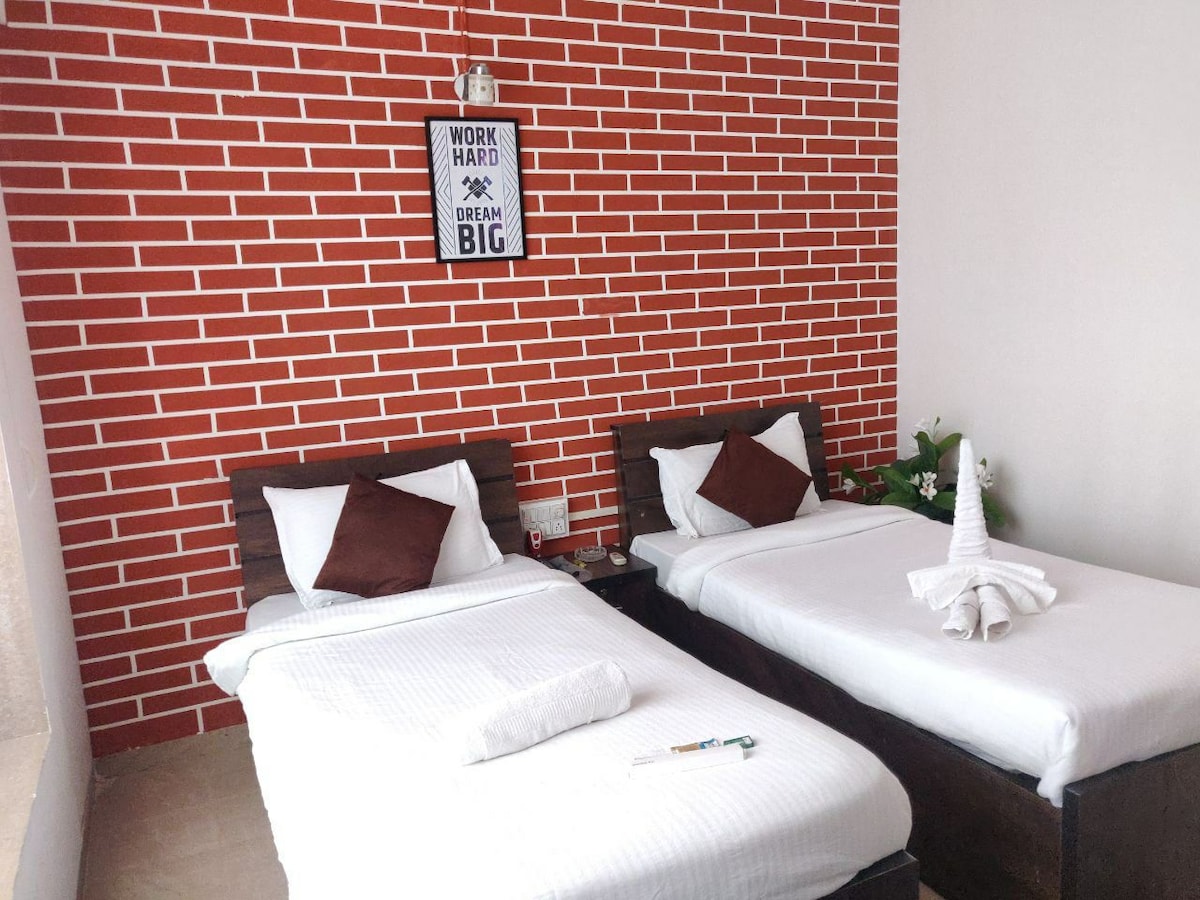 A room features two twin beds with white linens and brown wooden frames. A red brick wall serves as a backdrop, adorned with a motivational sign. A small plant is positioned beside the beds, contributing to the room's peaceful ambiance.