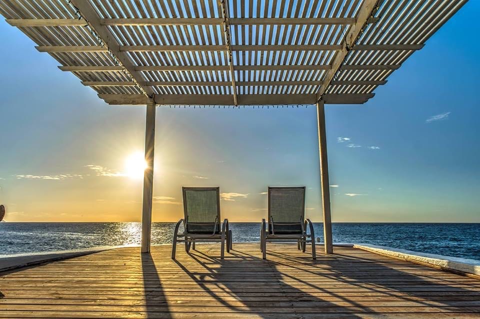 Two lounge chairs are positioned on a wooden deck beneath a slatted pergola, facing a stunning ocean view. The sun is setting on the horizon, casting warm light across the water, creating a tranquil and inviting atmosphere.