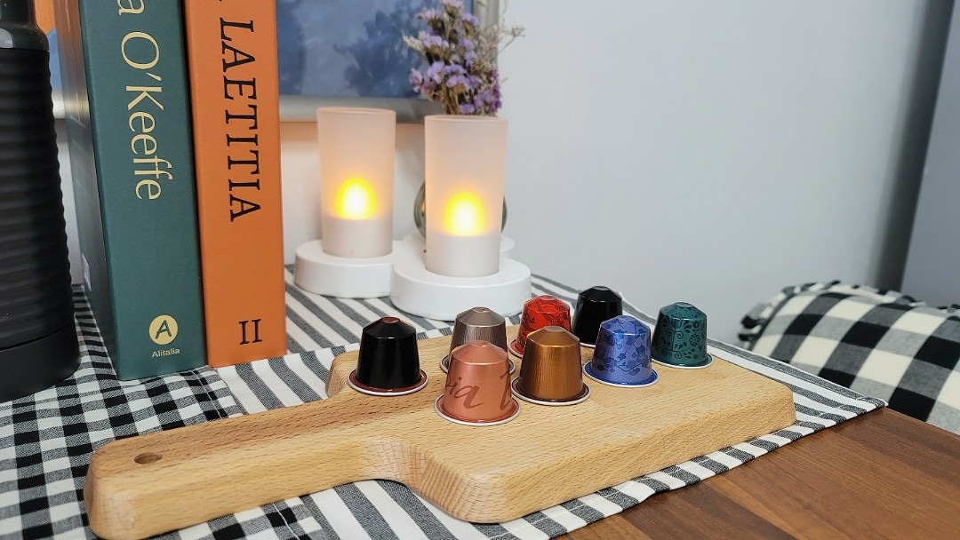 A wooden tray displays an array of colorful coffee capsules, arranged neatly alongside two softly glowing candles. Nearby, books are stacked in varying sizes, adding to the cozy ambiance. A checkered tablecloth completes the inviting setup.