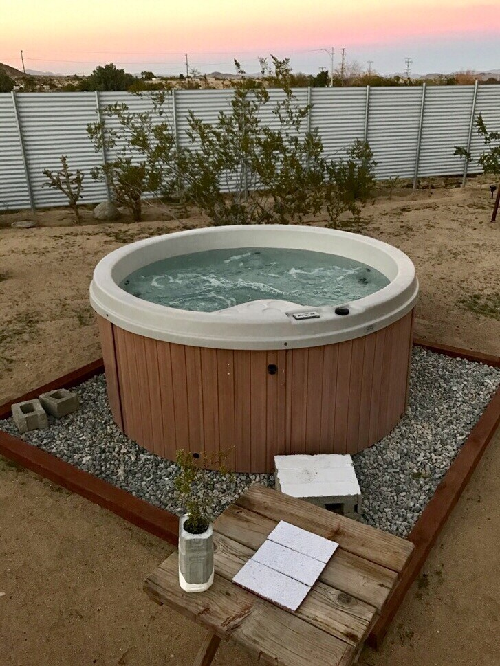 A circular hot tub, surrounded by gravel and wooden decking, is situated in an open outdoor space. A small wooden table with a potted plant rests nearby, complementing the serene natural landscape at sunset, with a soft glow in the sky.