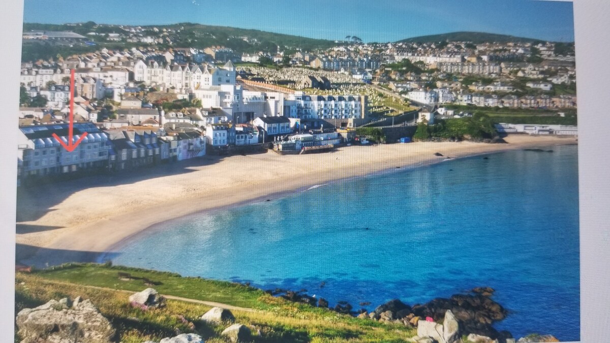 An aerial view highlights the waterfront of St Ives, showcasing golden sandy beaches meeting a calm blue sea. The flat's location is indicated on the left side of the image, surrounded by residential buildings along the shoreline and scenic hills in the background.