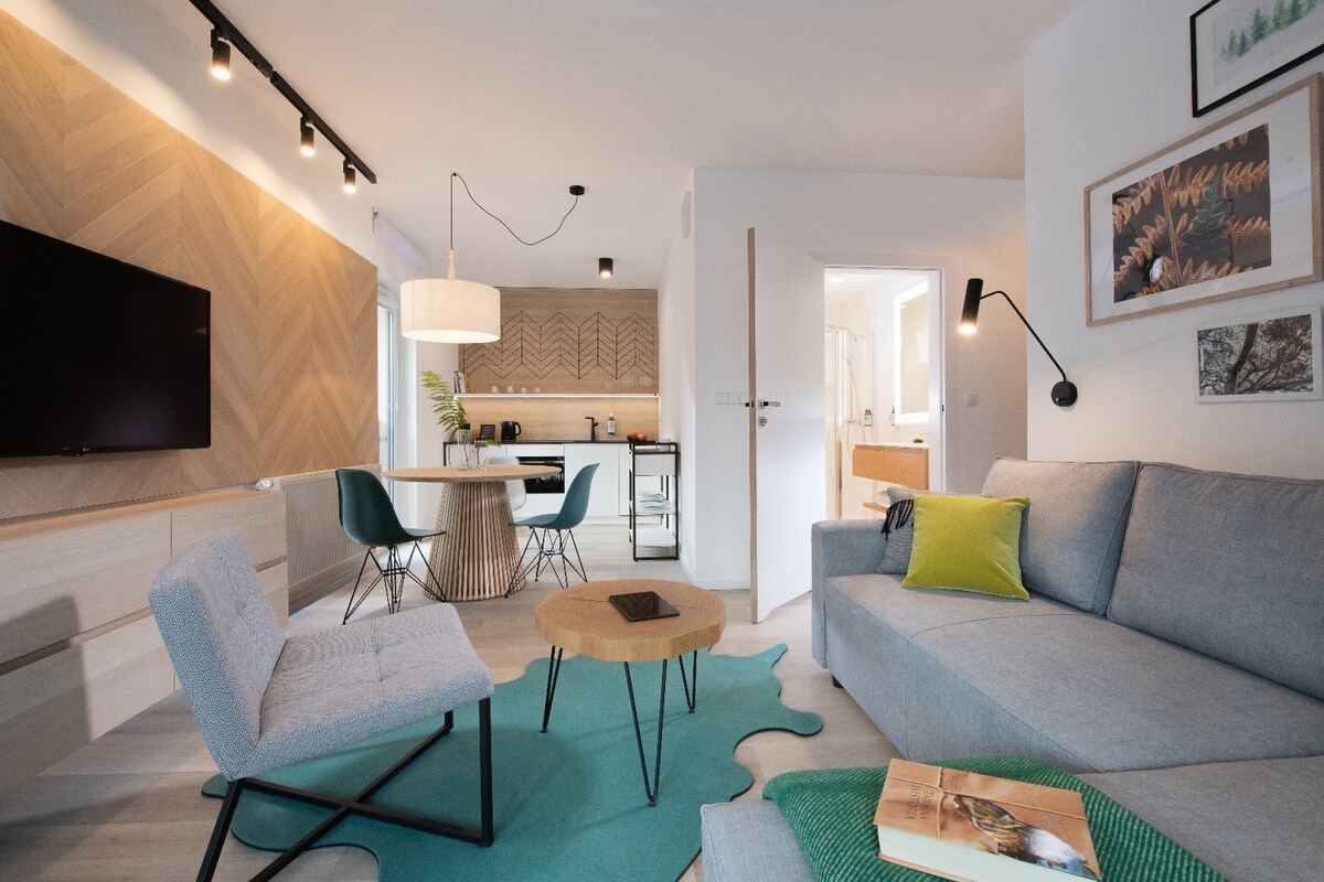 A spacious living area features a light grey sofa paired with an accent chair, set on a soft green rug. A wooden coffee table sits at the center, and a flat-screen TV is mounted on the textured wall. The kitchen area is visible in the background.