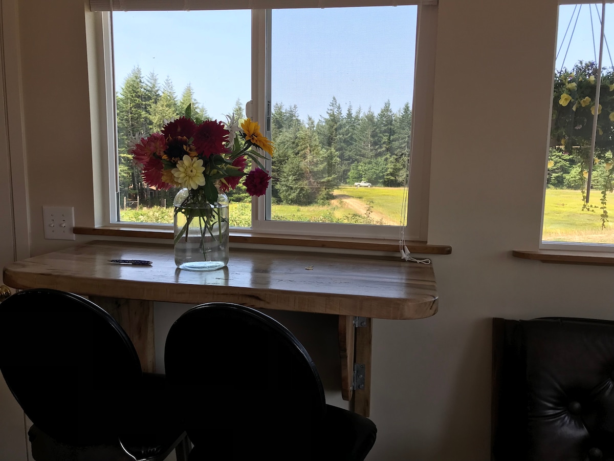 A small wooden table is positioned by a window, featuring a vase of colorful flowers. Two black chairs are placed beneath the table, providing a view of the surrounding greenery and timber outside.