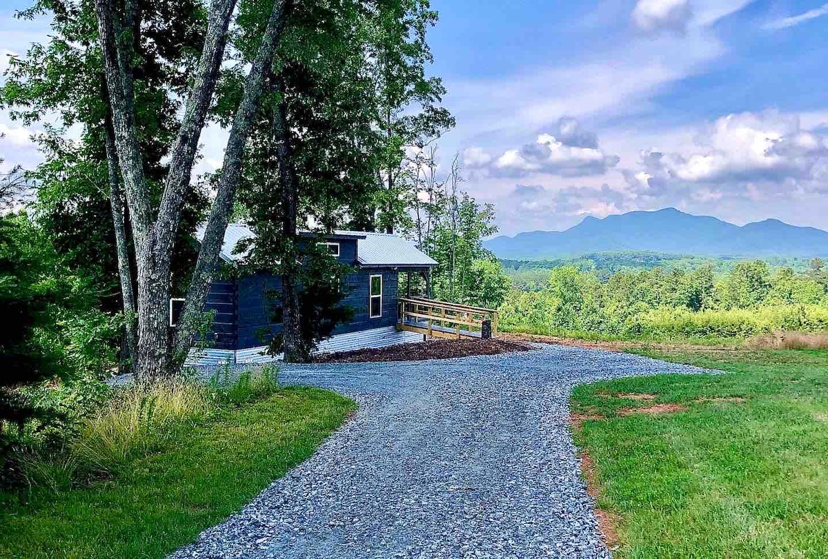 A gravel driveway leads to a secluded cabin surrounded by trees. Expansive views of the rolling hills and distant mountains are visible beyond the cabin. The landscape features lush greenery under a partly cloudy sky.