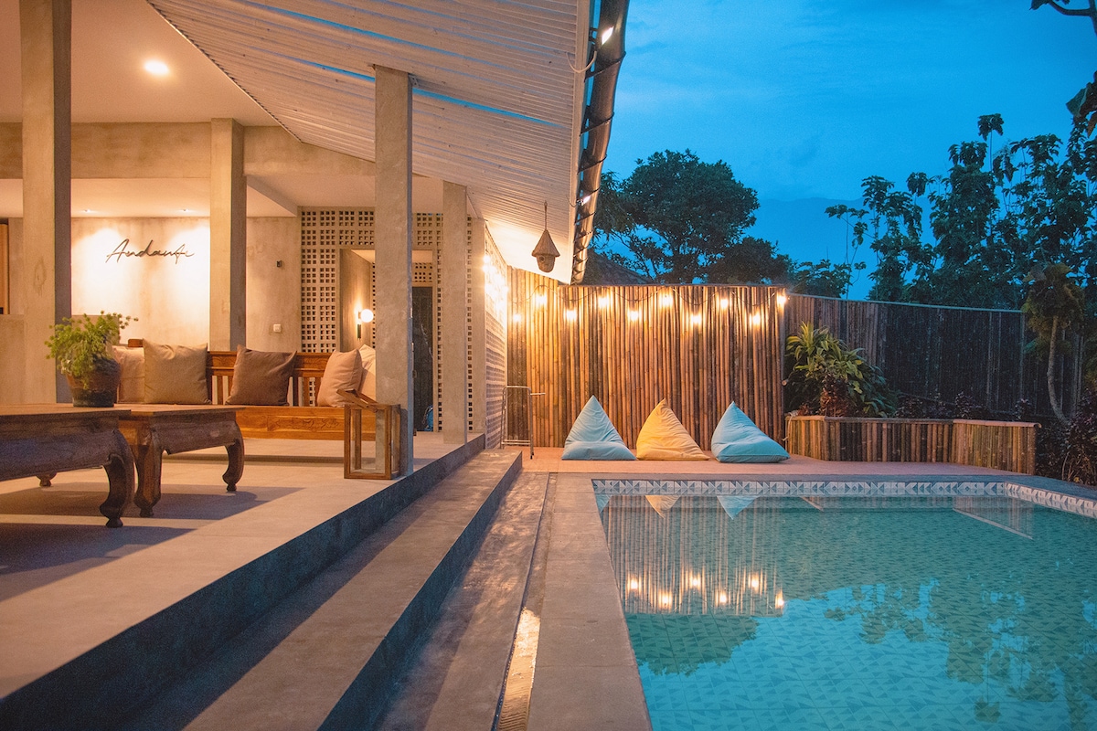 An outdoor space features a private swimming pool surrounded by sleek flooring. Comfortable seating areas are positioned nearby, with vibrant cushions placed on a wooden deck. Soft lighting enhances the inviting ambiance, creating a serene atmosphere as twilight approaches, complemented by greenery in the background.