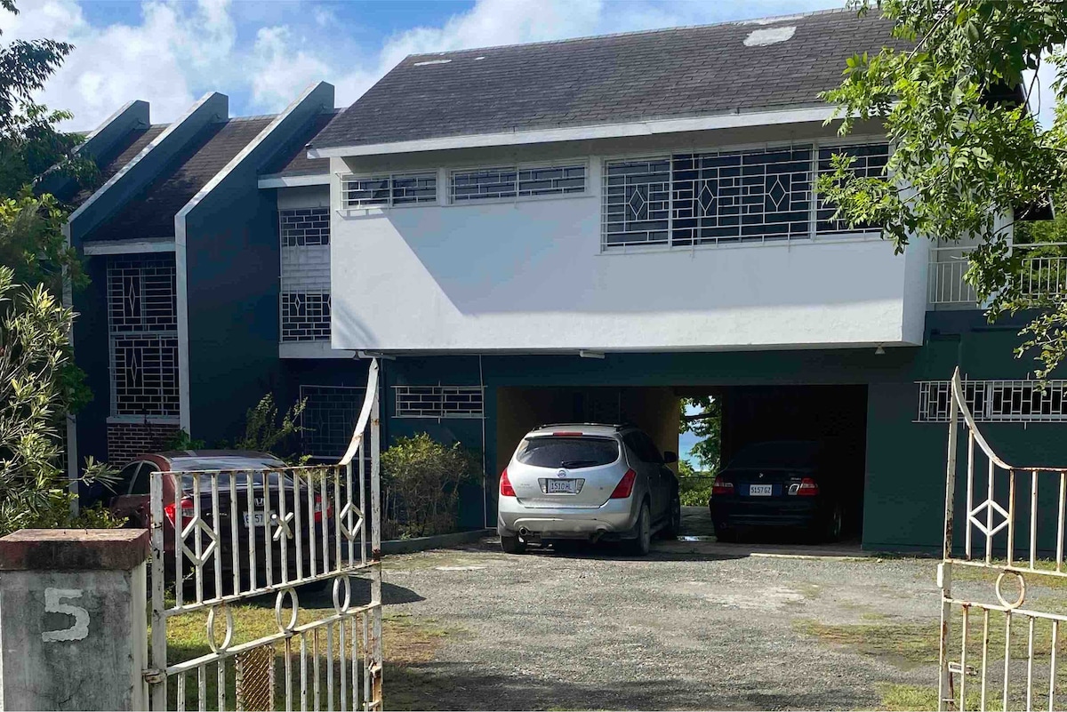 An exterior view of a two-level home features a covered parking area for multiple vehicles. The entrance is framed by a gated walkway, with greenery on either side. Large windows and a contrasting blue exterior add to the home's visual appeal.