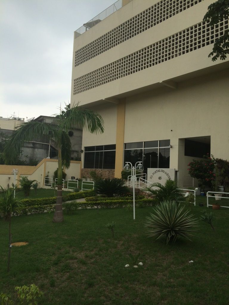 A modern building is showcased with a landscaped garden in the foreground. Palm trees and various shrubs surround the entrance, which features a gentle slope leading to the main door. Large windows provide a view of the interior, enhancing the inviting appearance.