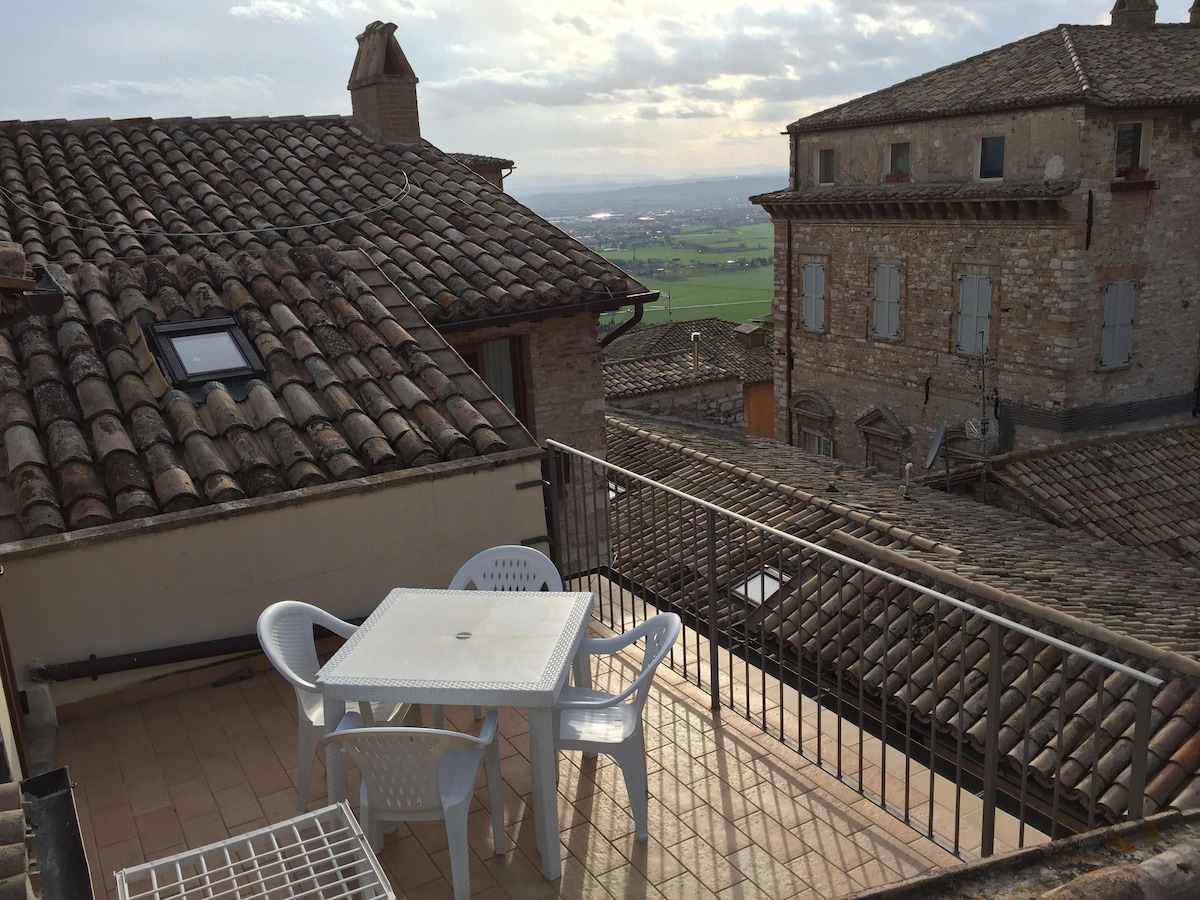 A rooftop terrace features a small white table and four chairs, offering a panoramic view of the surrounding landscape. The terrace is enclosed by a metal railing and overlooks traditional terracotta rooftops and stone buildings typical of the historic architecture in the area.