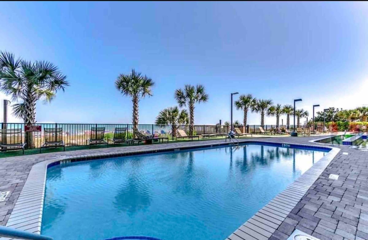 An outdoor pool area is framed by lush palm trees, creating a serene setting. The clear water reflects the blue sky, and lounge chairs are positioned around the pool. A fence separates the pool from the beach, which is visible in the background.