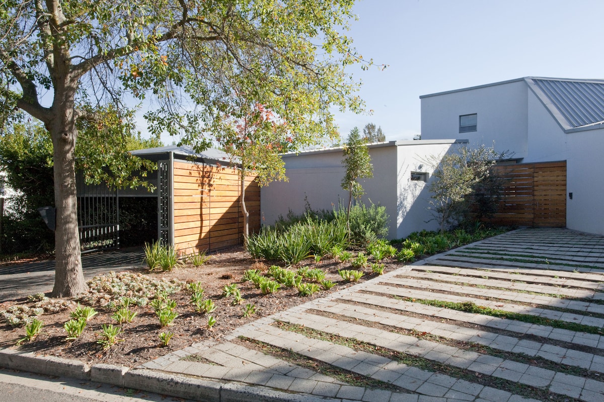 The exterior features a modern design with a combination of a wooden carport and a white facade. A landscaped area with low greenery complements the paved driveway, which leads to the entrance. A large tree provides shade, adding to the tranquil setting.