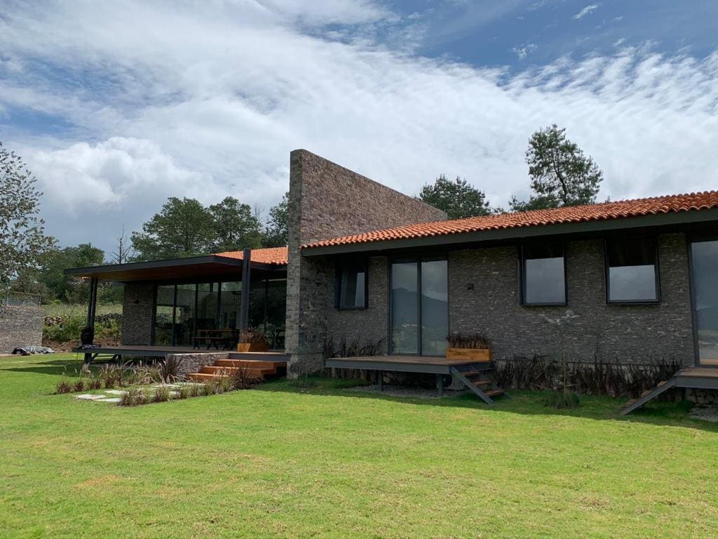 A modern structure is set in a grassy area, featuring a mix of stone and wood. Large glass doors provide views of the surrounding landscape. The roof is accented by orange tiles, and trees are visible in the background under a partly cloudy sky.