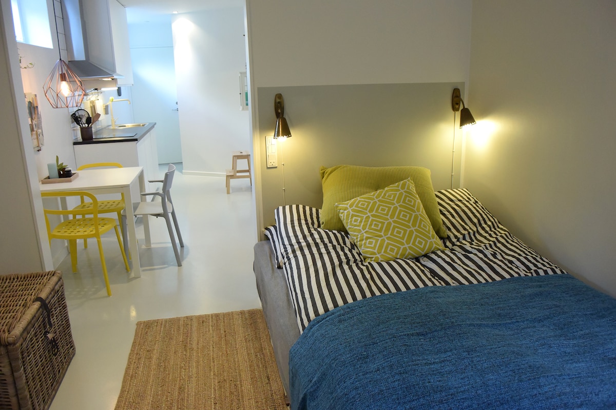 A compact living area includes a bed with striped linens and colorful pillows, accompanied by bedside lamps. A small dining table with yellow chairs is visible near a kitchenette, enhancing the light and functional design. A jute rug adds texture to the space.