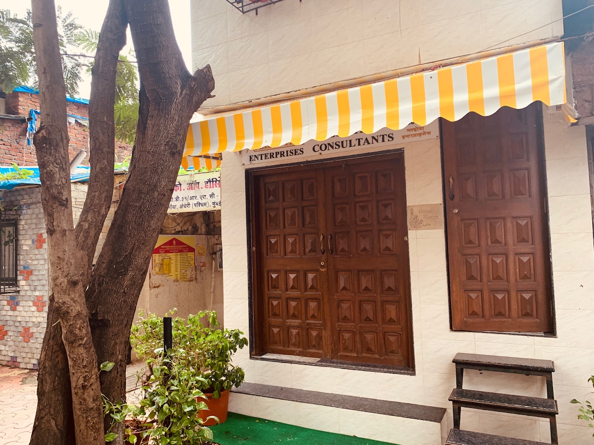 The exterior features a simple, white façade with a striped yellow and white awning above the entrance. Two wooden doors with decorative panels are framed by potted plants. A small set of steps leads to the door, with a tree providing shade nearby.