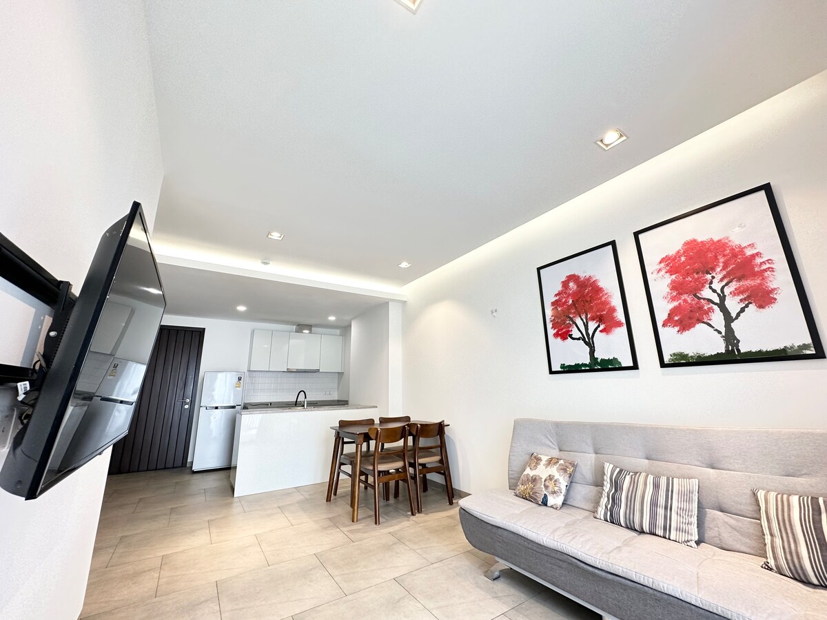 A modern living space features a gray sofa bed, wooden dining table with four chairs, and two framed artworks depicting red trees. Natural light highlights the tiled floor and kitchen area in the background, with a refrigerator and white cabinetry visible.