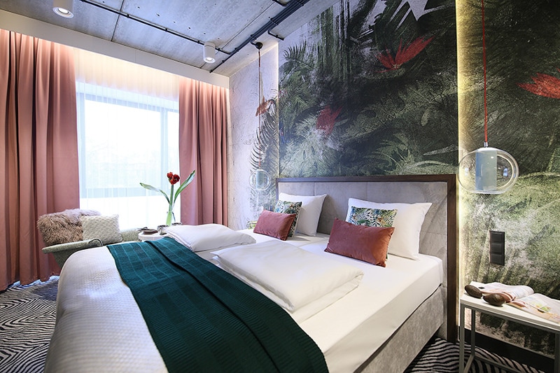 A modern bedroom features a spacious bed with white linens and colorful cushions. A green throw adds a pop of color. The walls display a vibrant jungle motif, complemented by large windows with sheer curtains. A chair with a soft throw can be seen beside the bed.