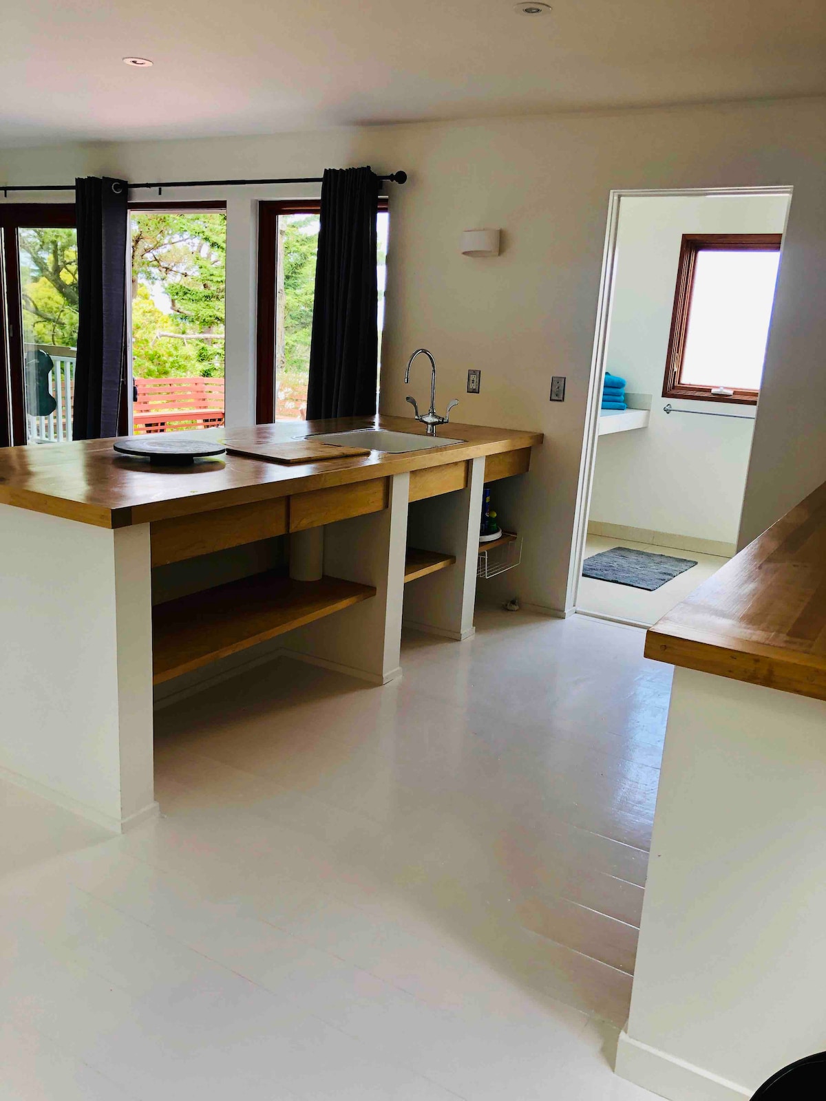 This spacious studio features a modern kitchenette with a wooden countertop and ample storage. Natural light floods in through large windows, while the bathroom is accessible through a door at the rear. Soft accents of greenery are visible outside, providing a refreshing ambiance.