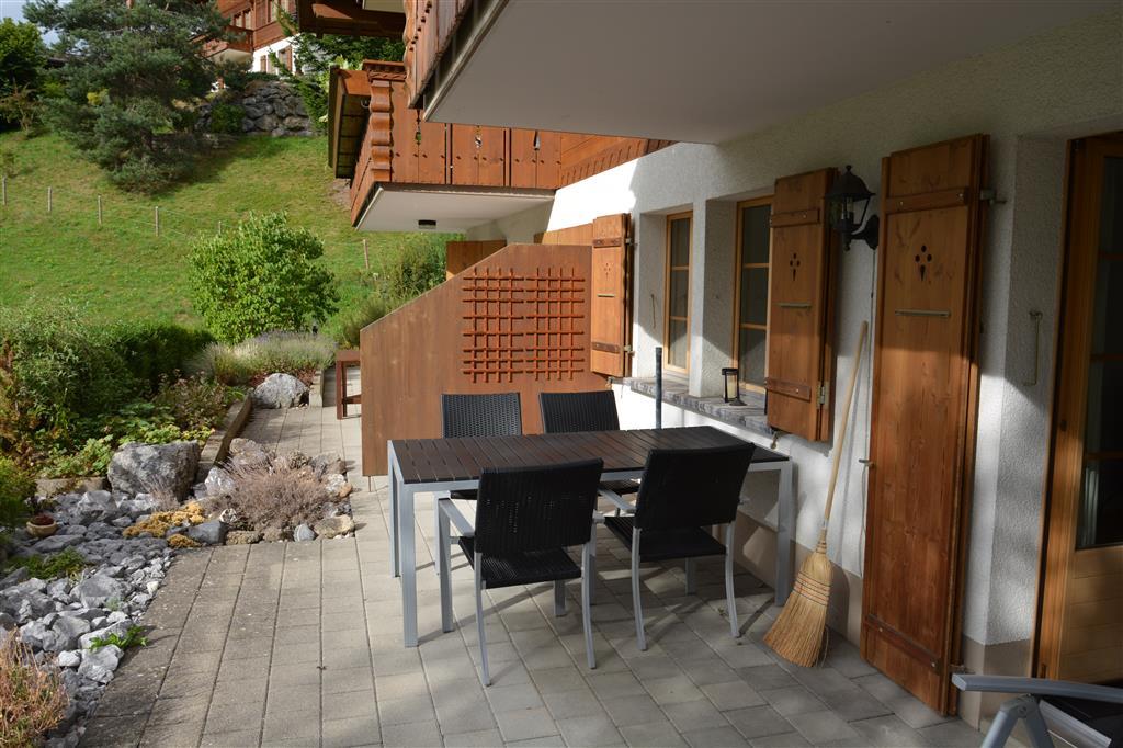 An outdoor terrace is featured, furnished with a table and four chairs. Wooden shutters frame the windows, while greenery and stone elements create a natural border. A broom leans against the wall, complementing the serene and functional outdoor space.