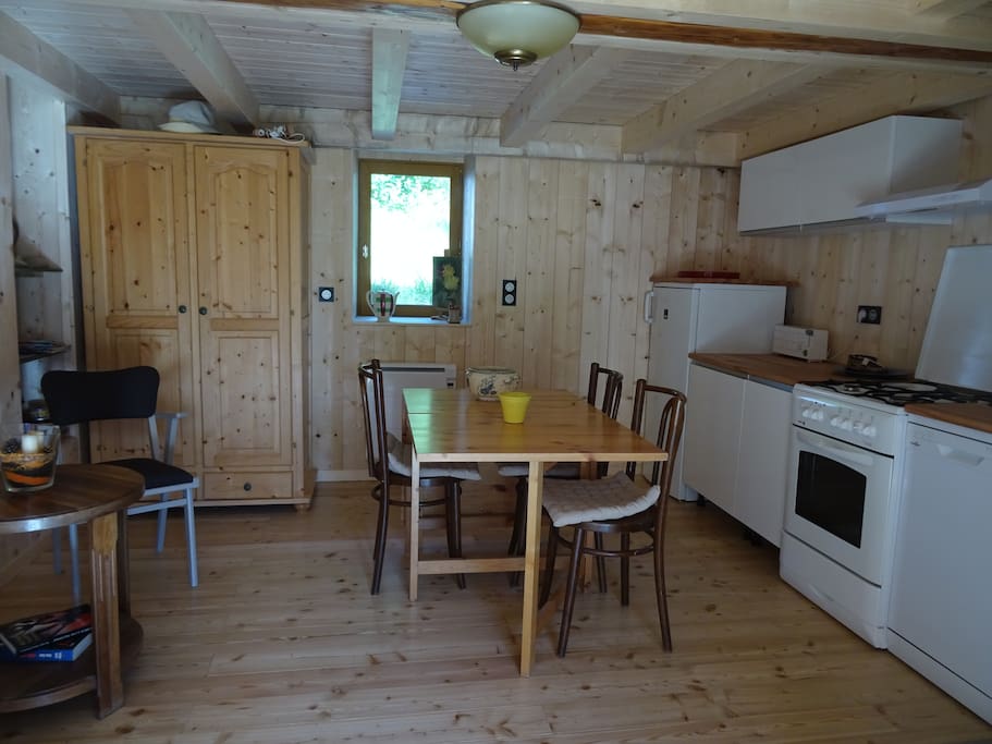 La pièce du rez de chaussée avec son coin cuisine : réfrigérateur, gazinière, lave-vaisselle, coin évier, grill-pain, bouilloire, cafetière Senséo, coin repas et petit coin repos à côté du poêle, armoire penderie et étagères