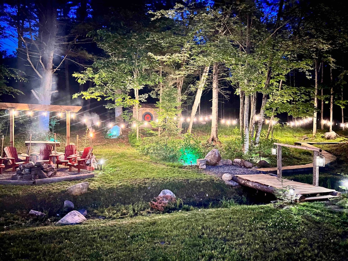 A cozy outdoor seating area is surrounded by trees, featuring a circular arrangement of wooden chairs around a fire pit. A wooden bridge crosses a small stream, while string lights illuminate the area, creating a warm ambiance amidst the natural surroundings.
