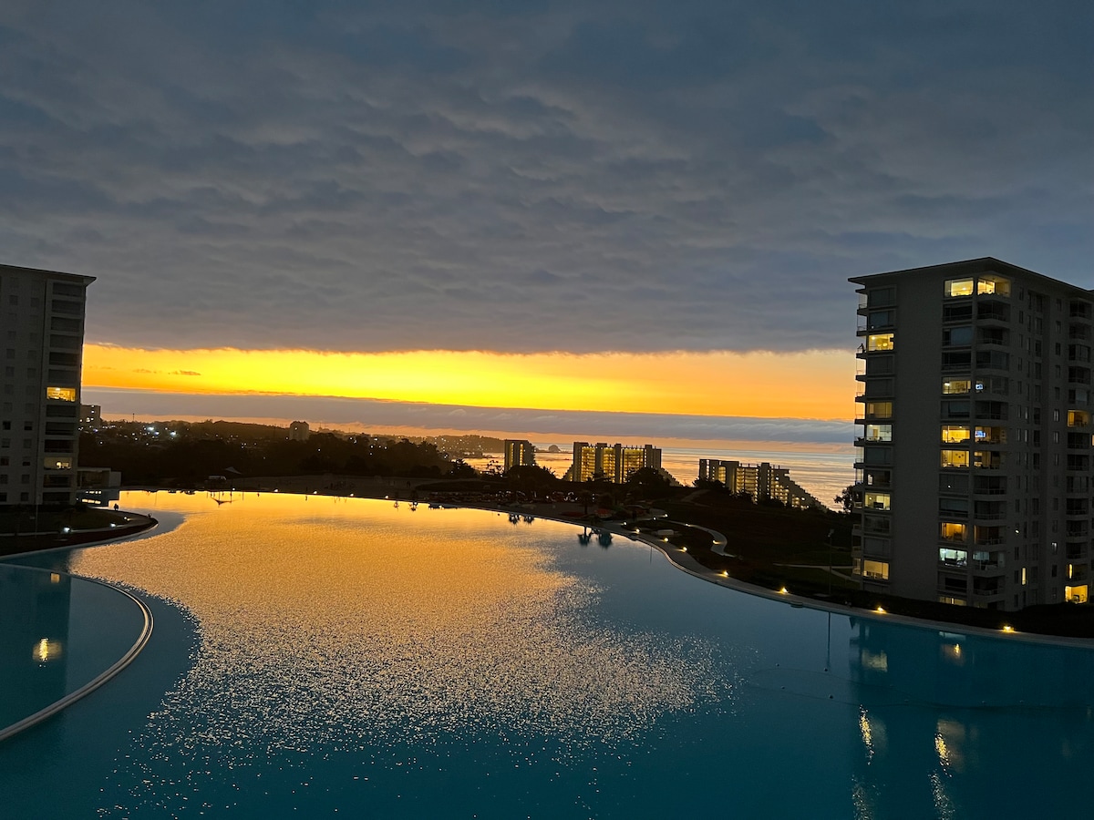 A serene view captures the sunset over a large, tranquil pool, reflecting vibrant hues of orange and yellow. Towering buildings flank the pool, with warm lights glowing from their windows. Soft clouds hang above, enhancing the calmness of the scene.