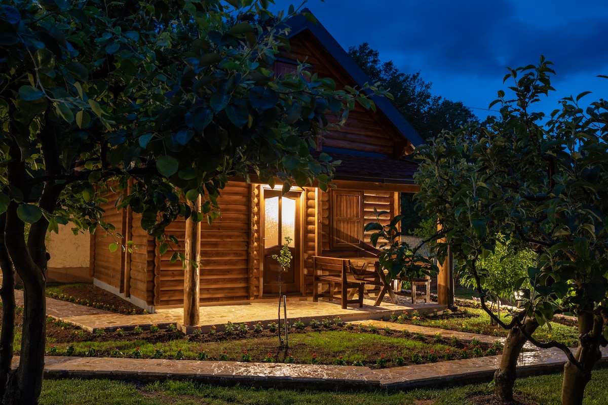 A charming wooden chalet is illuminated by warm light from the entrance, surrounded by lush greenery. A stone path leads to the door, with a seating area visible on the porch. Fruit trees are positioned around the chalet, enhancing the peaceful outdoor setting.