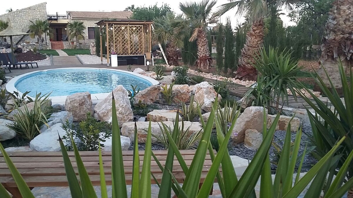 A serene outdoor space features a sparkling swimming pool surrounded by smooth stones and lush greenery. Palms and other plants enhance the natural ambiance, while a wooden bench provides a spot for relaxation. A pergola offers shade in the background, complementing the tranquil environment.
