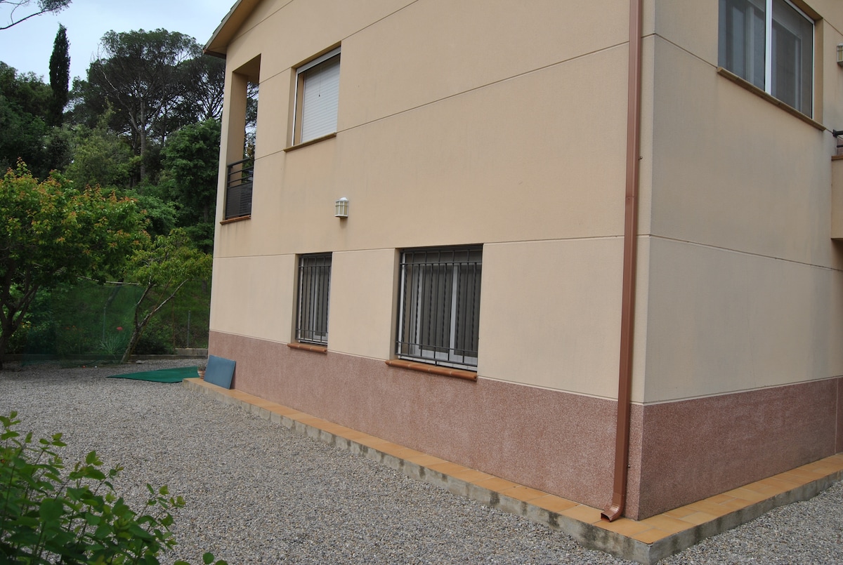 The exterior of the ground floor apartment is shown, featuring a peach-colored façade with large windows framed by bars. A gravel area surrounds the building, providing space for outdoor activities. Lush greenery is visible in the background, contributing to the setting’s natural environment.