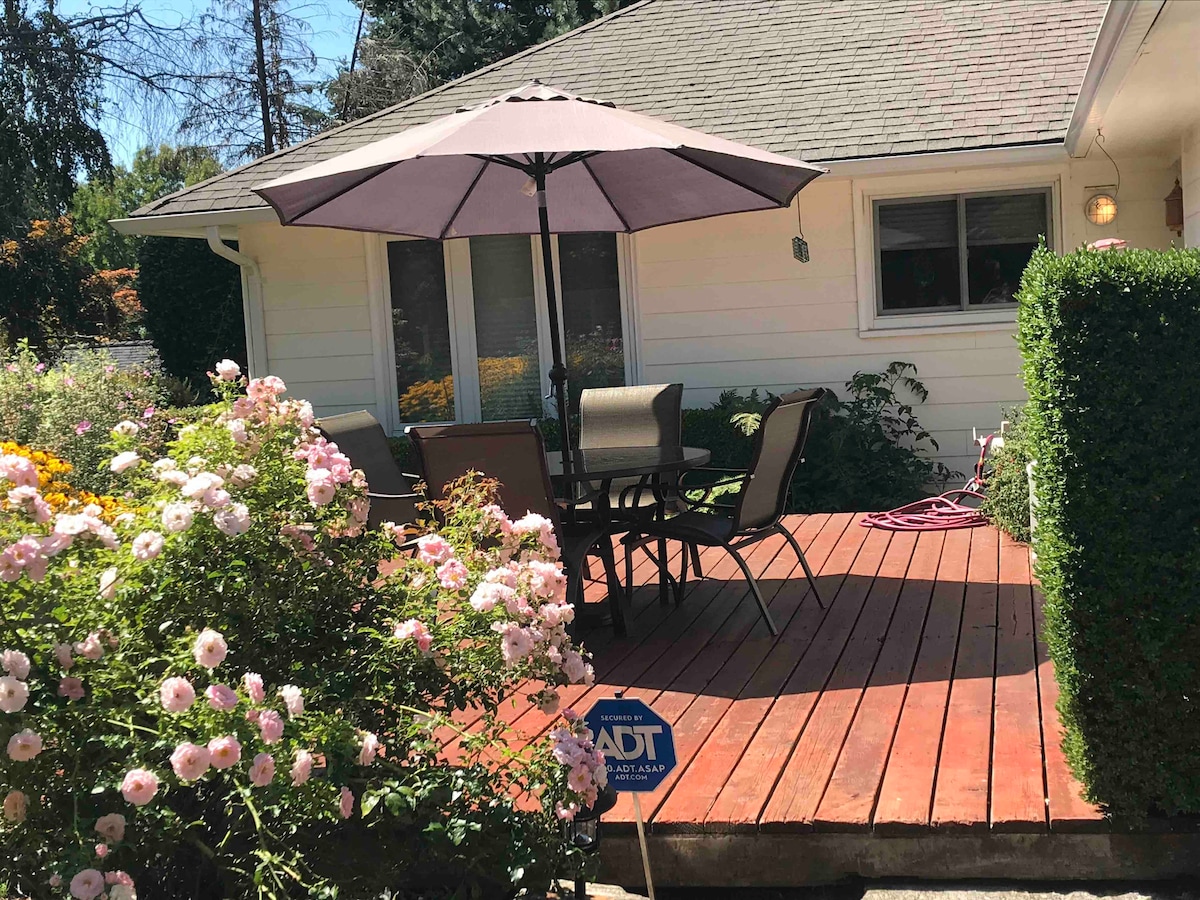 An outdoor deck is shown, featuring a round umbrella over a set of six chairs arranged around a wooden table. Surrounding the deck, vibrant rose bushes add color to the setting, while sunlight brightens the area.