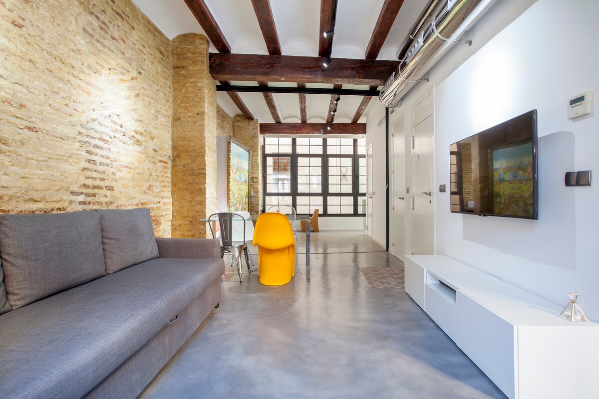 A spacious living area showcases exposed stone walls and wooden beams. A large gray sofa is placed adjacent to a bright yellow chair and a minimalist white entertainment unit. Natural light floods in through the expansive reflective glass window, enhancing the open feel of the room.