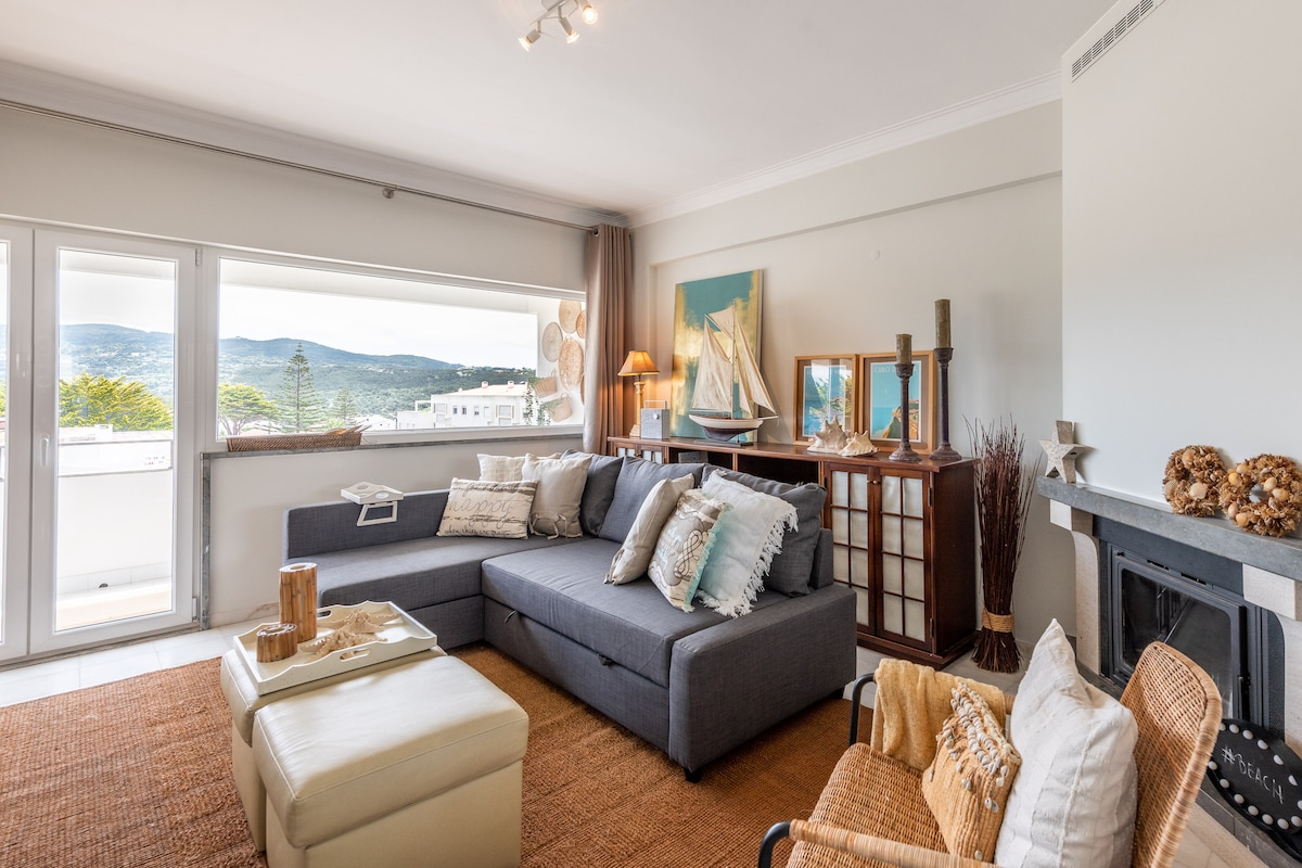 A modern living area features a spacious sectional sofa complemented by decorative cushions. Natural light floods in through a large window with scenic views of the surrounding hills. A stylish wooden cabinet and an art piece add character to the room, enhancing its inviting ambiance.