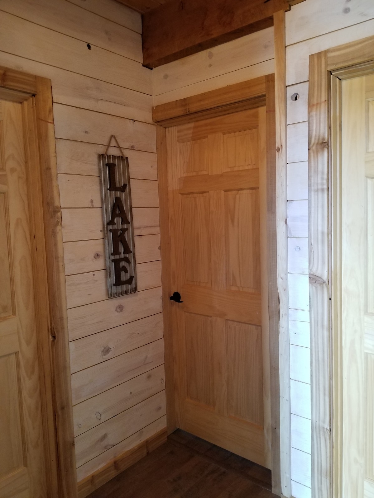 The image features a hallway with natural wood paneling on the walls. Two wooden doors are visible, with one labeled 'LAKE' on a sign hung nearby. The doors have simple handles, and the flooring is also wood, contributing to a rustic aesthetic.