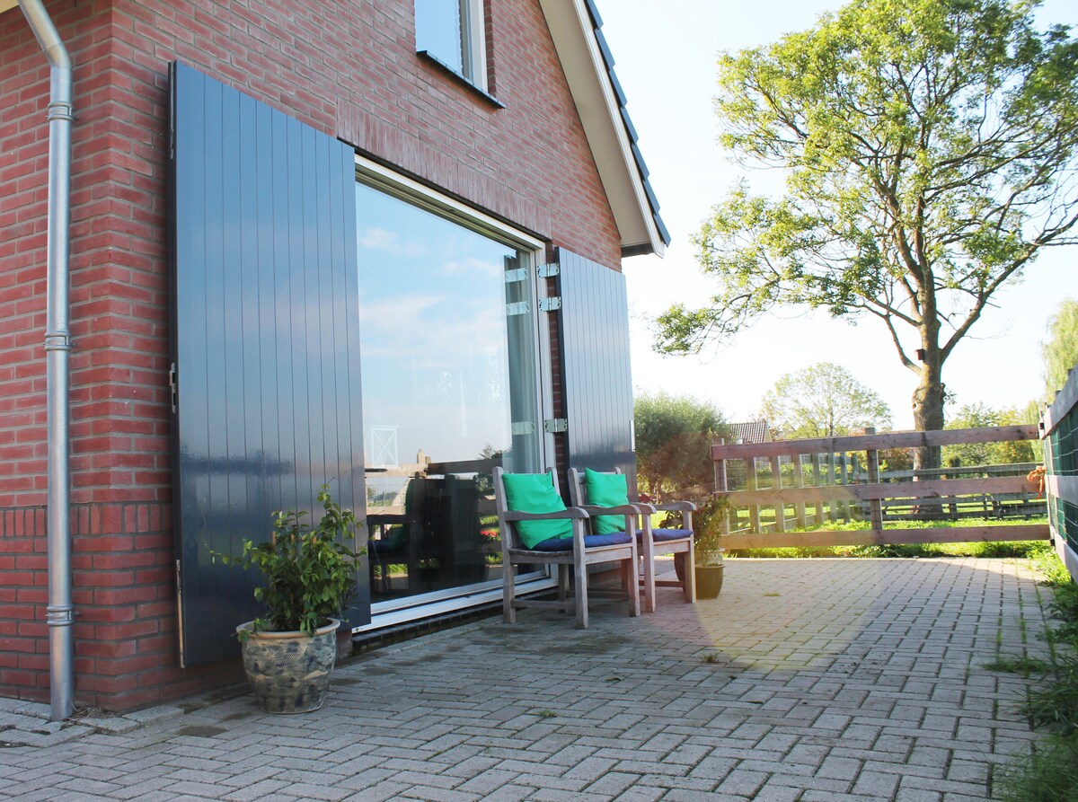 A patio area features a set of wooden chairs with green cushions, positioned beside large glass doors that open to the interior. A potted plant sits on the ground. The surrounding landscape includes trees and greenery, creating a serene outdoor setting.