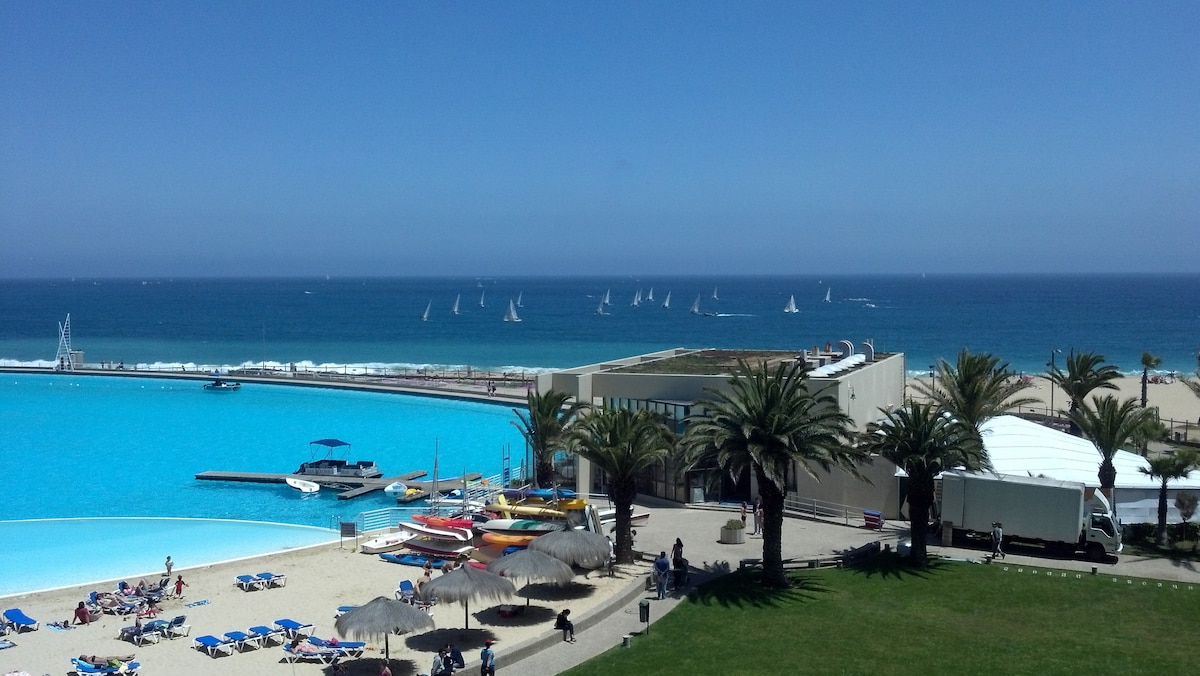 A clear view of a tranquil lagoon with a vibrant turquoise hue, bordered by sandy areas and palm trees. In the background, sailboats can be seen gliding across the open sea, while lounge chairs and shaded umbrellas offer relaxation options near the water.