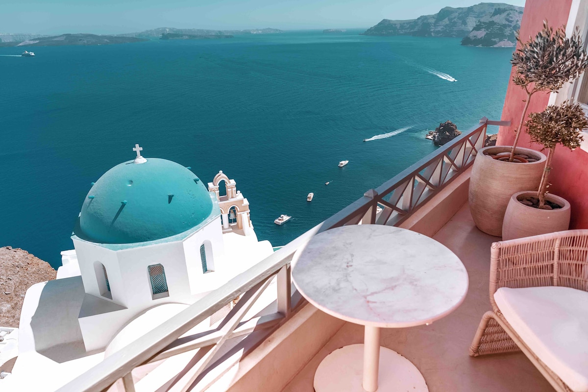 A balcony area is visible with a round marble table and a woven chair, overlooking the Aegean Sea. The striking blue dome of a nearby church adds a focal point, while boats can be seen on the water. The cliffs' colors enhance the coastal view.