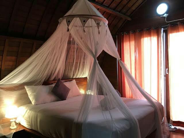 A spacious bed is draped with a delicate canopy, creating a peaceful sleeping area. Soft lighting is provided by a lamp on the bedside, while orange curtains filter natural light from the window, adding warmth to the wooden interior.