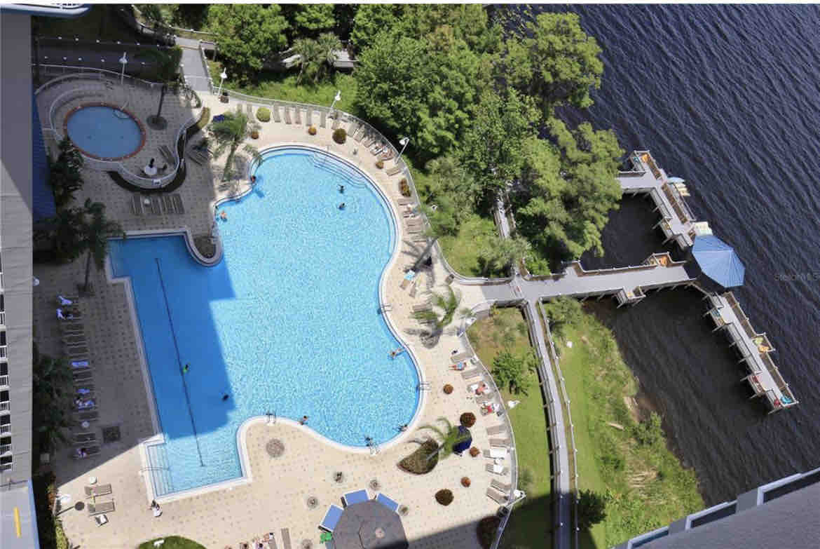 An aerial view reveals a large, inviting pool surrounded by palm trees and lounge chairs. A small hot tub is visible nearby, and a wooden dock extends towards the water. The area is lushly landscaped, providing a natural backdrop to the serene setting.