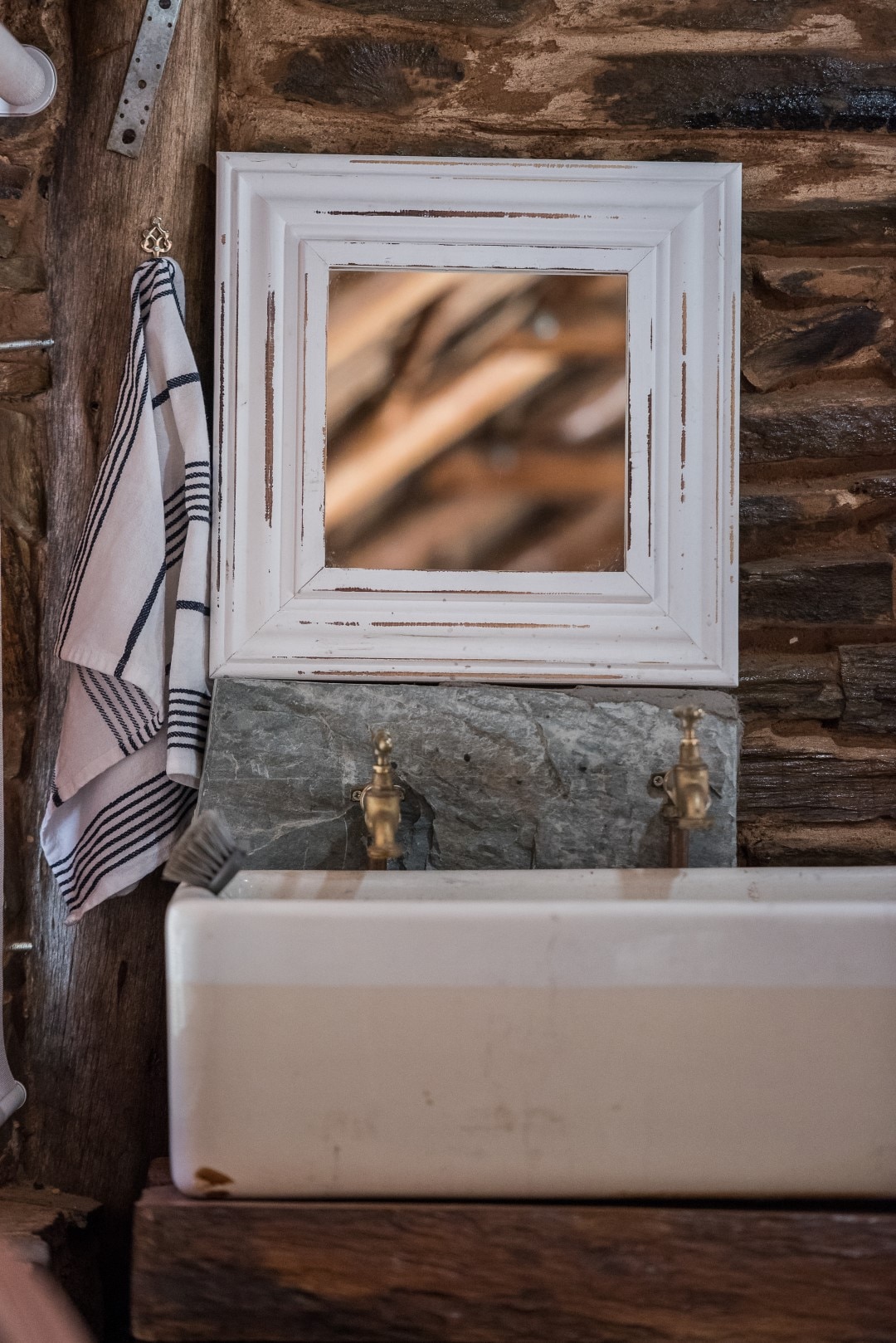 The bathroom features a rustic stone wall, complemented by a white-framed mirror above a vintage-style wash basin. Two brass faucets are visible, and a striped towel hangs nearby, adding a touch of practicality to the serene setting.