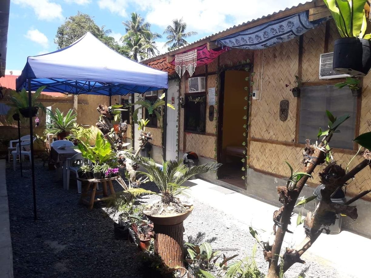 A garden area is surrounded by bamboo structures, featuring a blue canopy providing shade. Potted plants and greenery enhance the space, while a stone pathway guides toward individual entrances. Comfortable seating arrangements can be noted with white chairs positioned around small tables.