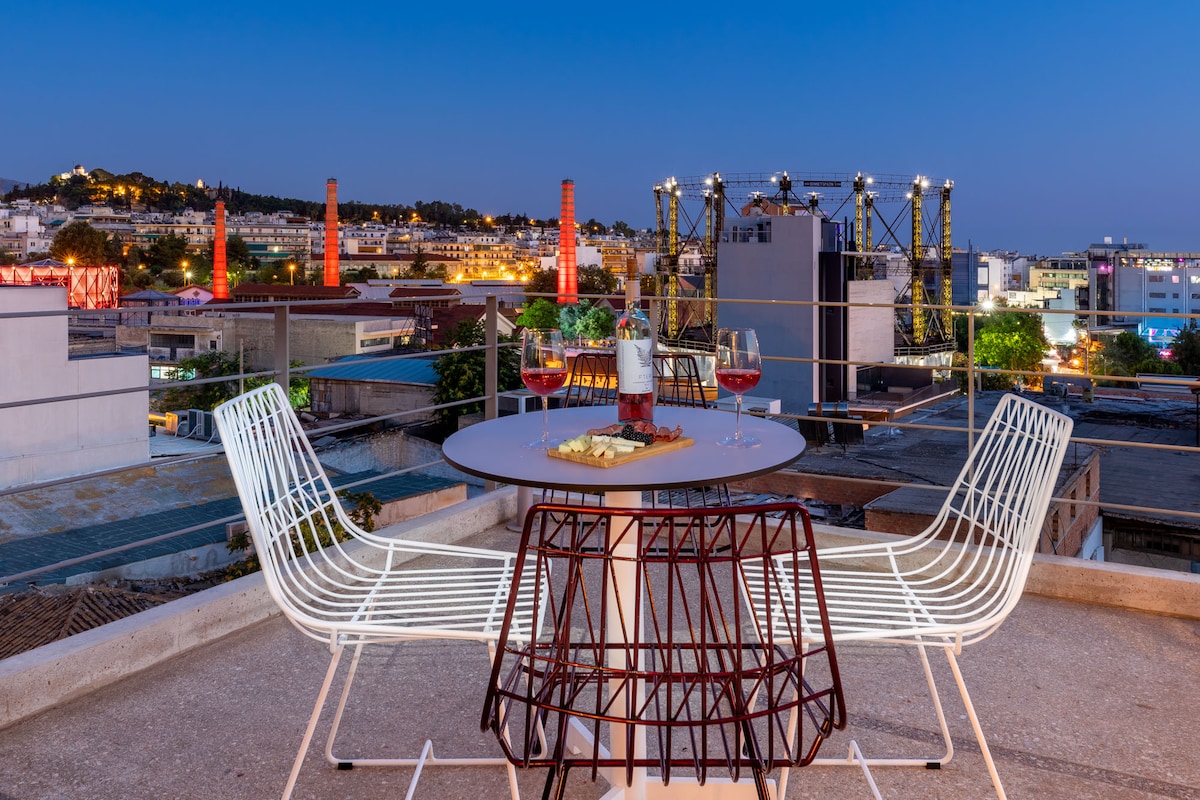 A private balcony showcases a round table surrounded by white wire chairs, set against a backdrop of the illuminated Athens skyline at dusk. The scene features colorful lights from nearby buildings and an expansive view of the rooftops, creating a vibrant urban atmosphere.